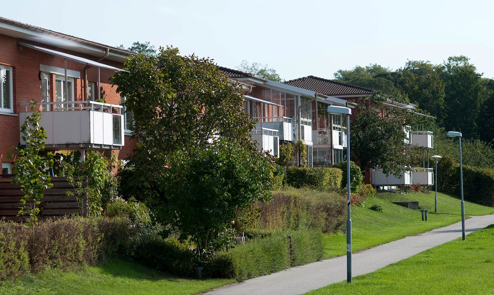 En rad moderna hus med röda tegelfasader och vita balkonger, omgivna av grönska och en gångväg med gatlyktor. Himlen är klar.