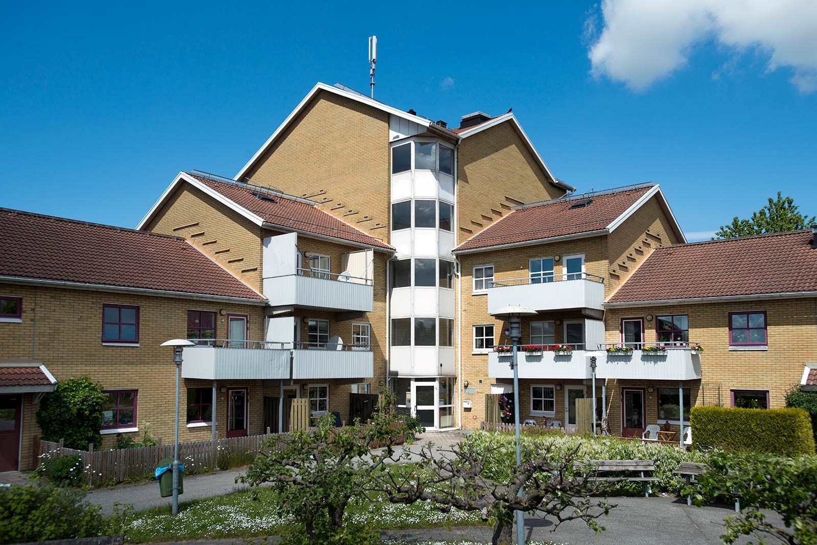 En ljusgul byggnad under en klarblå himmel. Byggnaden har tre våningar med en central del som sträcker sig högre än de omgivande flyglarna. Varje lägenhet har en balkong, och det finns gröna träd och buskar runt byggnaden,