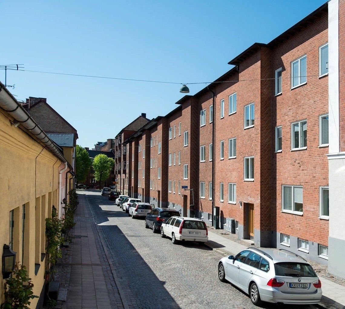 En lugn och solig stadsgata. På ena sidan syns röda tegelbyggnader med jämnt fördelade fönster. Til