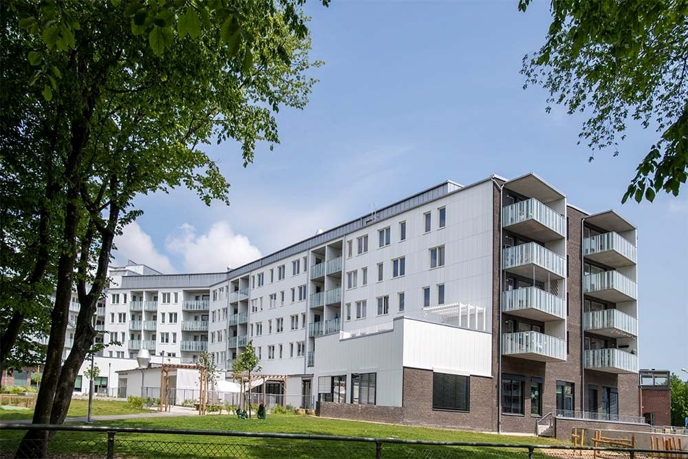 Bilden visar en modern vit byggnad med flera våningar, omgiven av gröna träd och en klar blå himmel
