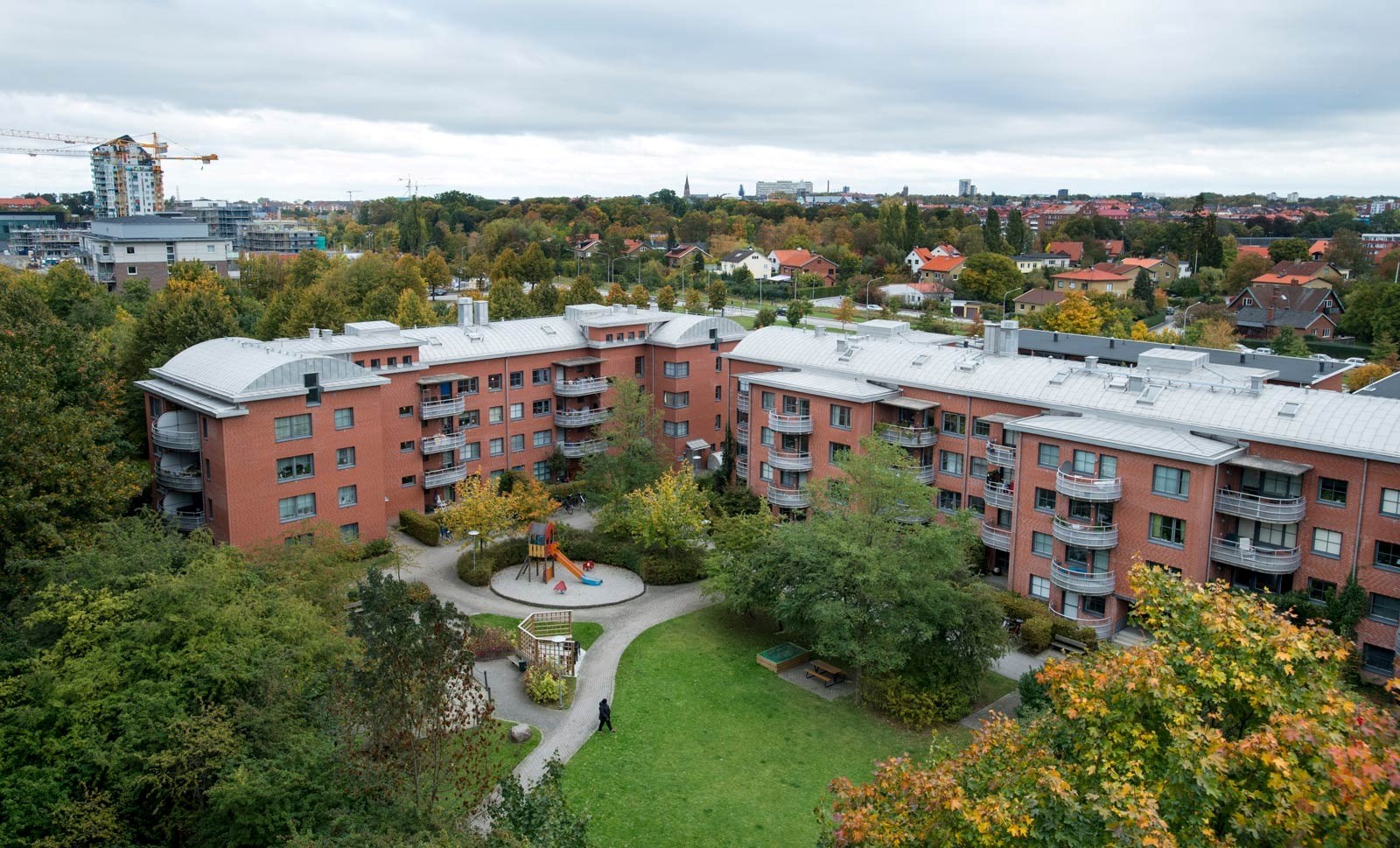 En utsikt över ett bostadsområde med röda tegelbyggnader, en lekplats, och träd med höstfärger. En 