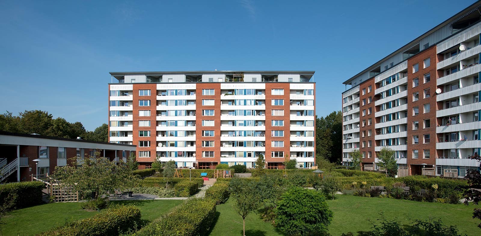 En bostadsbyggnad omgiven av gröna ytor under en klar himmel, med flera våningar och balkonger som ger en känsla av öppet utrymme och gemenskap.