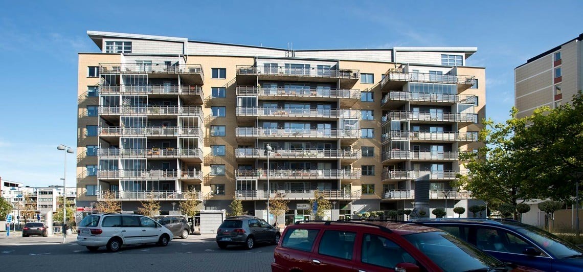 En modern flervåningsbyggnad med balkonger, omgiven av parkerade bilar under en klar himmel. Fasade