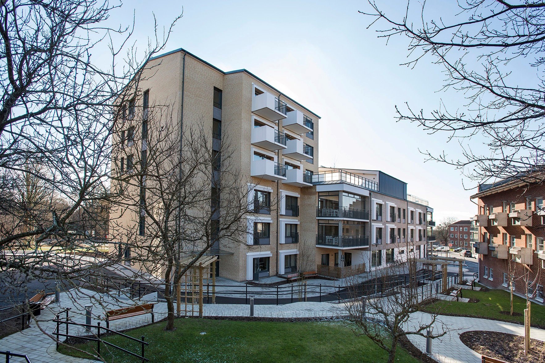 En modern bostadsbyggnad omgiven av en välplanerad utomhusmiljö. Byggnaden har flera balkonger och fönster. En stig leder fram till byggnaden, omgiven av gräs och andra landskapselement.