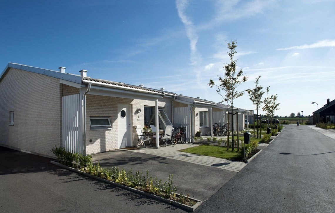 En rad moderna enfamiljshus med en ren och minimalistisk design, belägna längs en asfalterad gata. Varje hus är vitt med en tegelfasad och har en liten, välskött framsida.