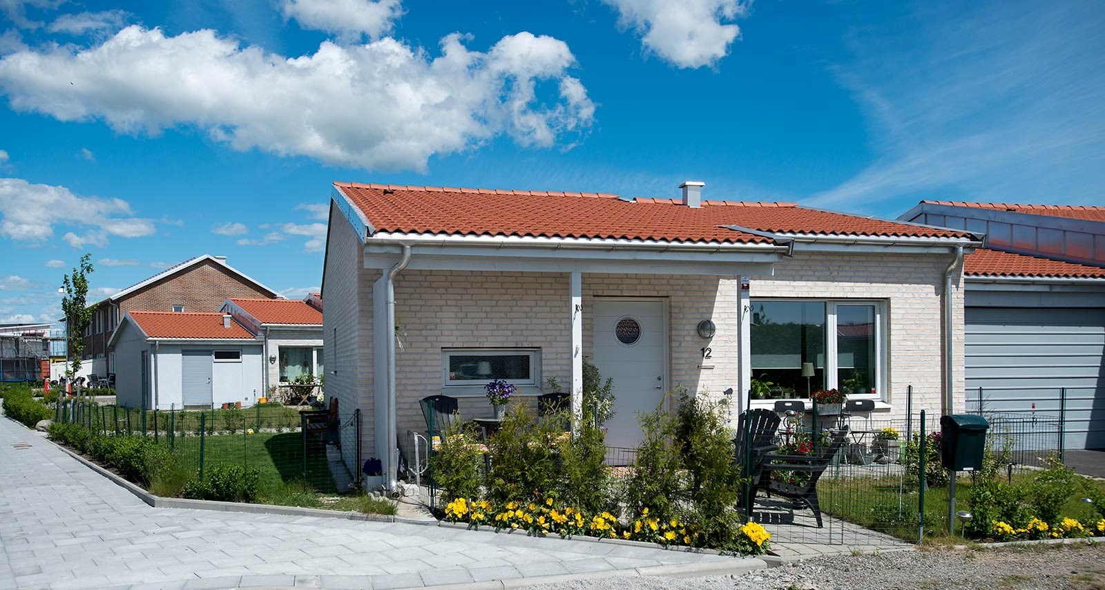 Ett modernt enfamiljshus i en våning med en ljusfärgad tegelfasad och ett rött tegeltak mot en klarblå himmel med några moln. Framför huset finns en välskött trädgård med olika växter och blommor, främst gula, som omger en prydlig gräsmatta. En stenlagd gång leder till husets entré.