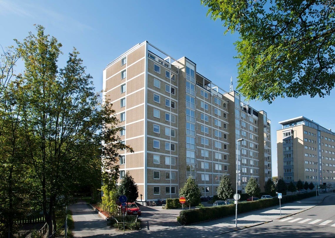 En stor bostadsbyggnad omgiven av grönska och en klarblå himmel. Byggnaden är ljusbrun med många fönster som reflekterar himlen. På båda sidor om byggnaden finns träd. En väg leder upp till entrén där några cyklar är parkerade.