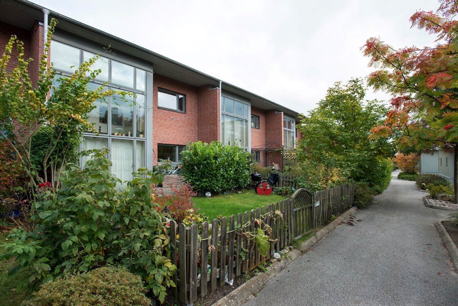 Ett bostadsområde med röda tegelhus och en trästaket längs en grusväg. Träden börjar få höstfärger och himlen är ljus.