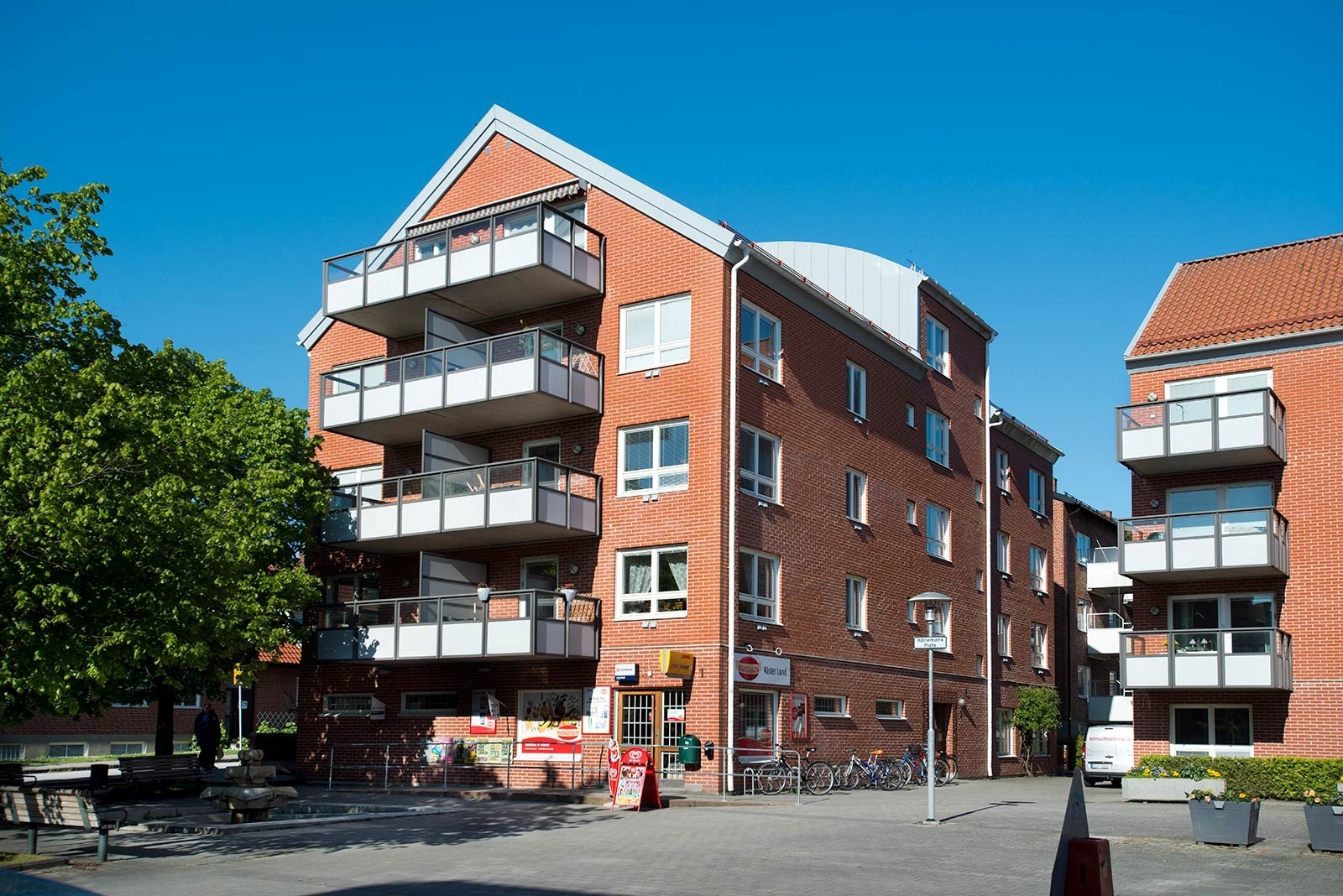 Ett flerfamiljshus i rött tegel placerat vid ett litet torg. På bottenplan finns ett livs.