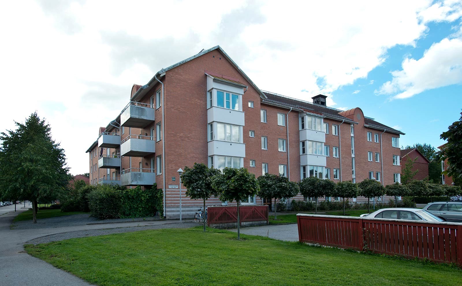 Ett flerfamiljshus med tegelfasad och vita balkonger. I förgrunden syns en parkeringsplats
