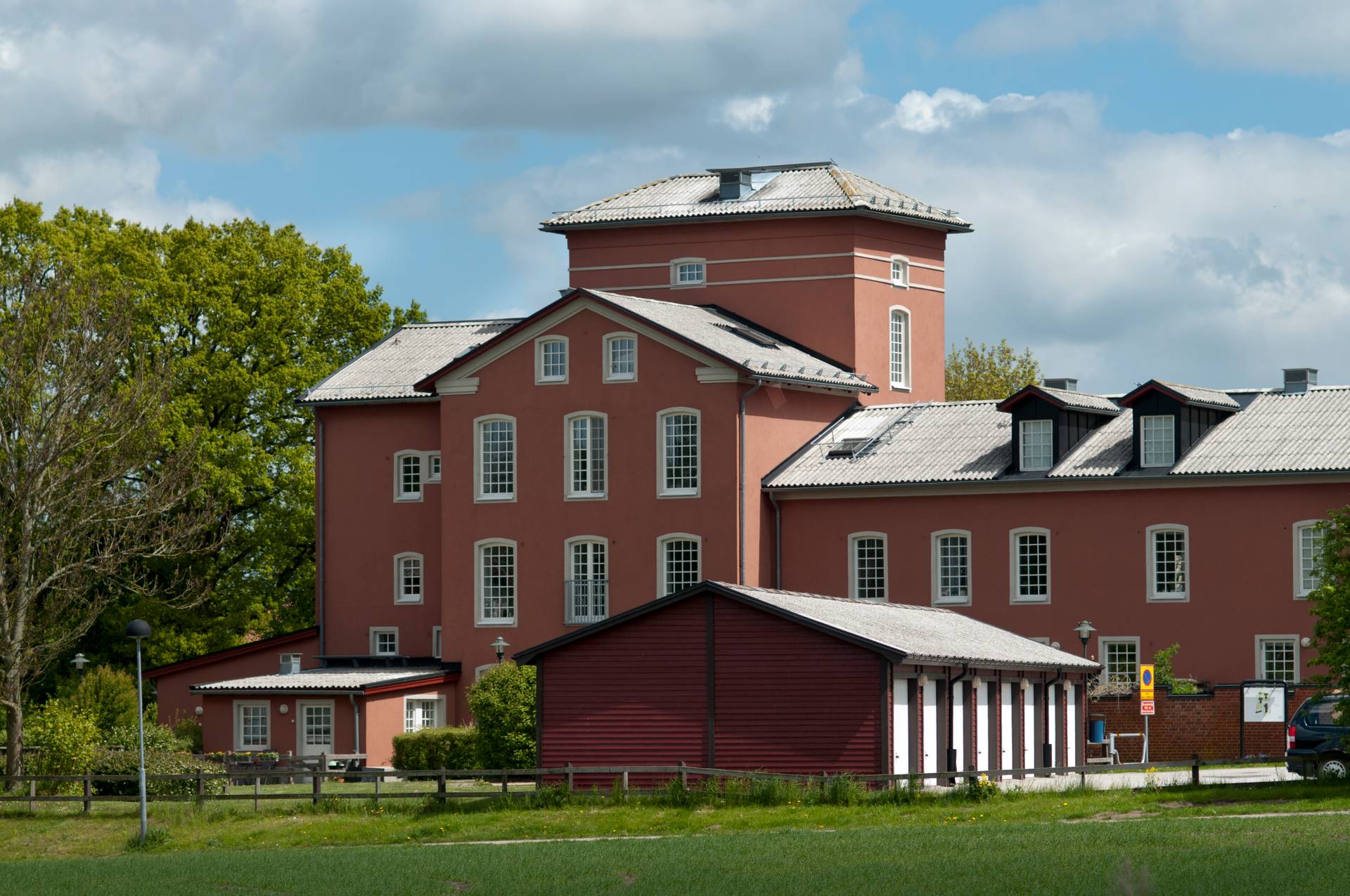 Ett stort, flervåningshus med en framträdande röd fasad och vita detaljer runt fönster och takkanter. Byggnaden har en symmetrisk design med en central del som är högre än de omgivande flyglarna, och har ett sadeltak. Framför huvudbyggnaden finns en mindre röd byggnad med vita dörrar och ett angränsande vitt staket. Miljön inkluderar grönt gräs i förgrunden och träd i olika gröna nyanser bakom byggnaderna under en delvis molnig himmel.