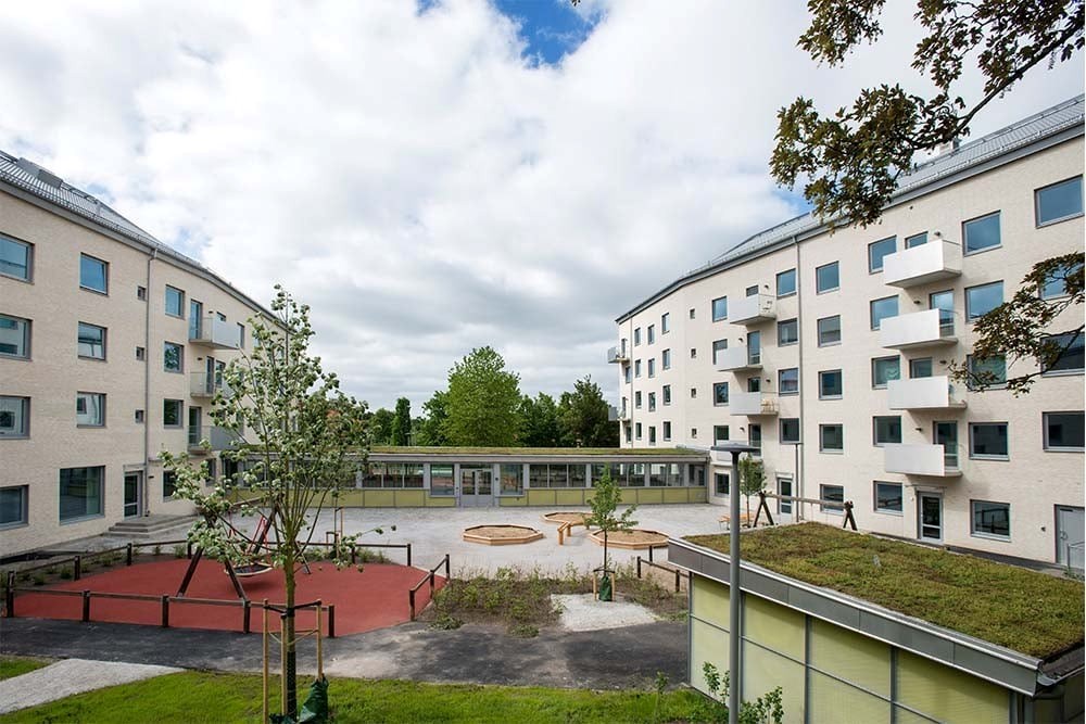 Två stora huskroppar med ljust tegel. Mellan husen är en innergård med lekplats och förrådsbyggnad med grönt tak