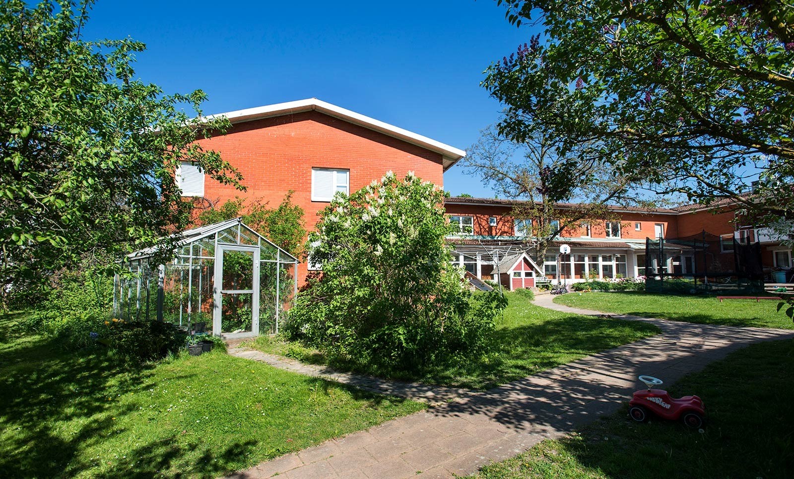 En solig trädgård med gröna träd och gräs, en röd tegelbyggnad i bakgrunden. En växthusstruktur och en liten röd leksaksbil på marken. Atmosfären är fridfull och lugn.