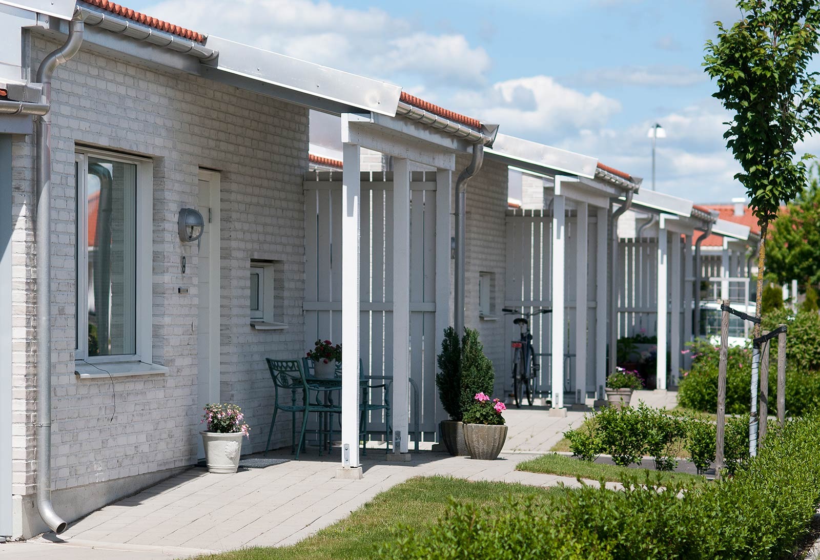 En rad moderna, enplans radhus. Varje hus har en vit tegelfasad, stora fönster med mörka ramar och ett lutande tak med röda tegelpannor. Husen är utrustade med vita markiser ovanför fönster och dörrar. Framför ett av husen finns en liten uteplats med ett runt bord och två stolar. Dekorativa växter i krukor syns nära ingången till detta hus. En prydligt stensatt gångväg löper framför husen.