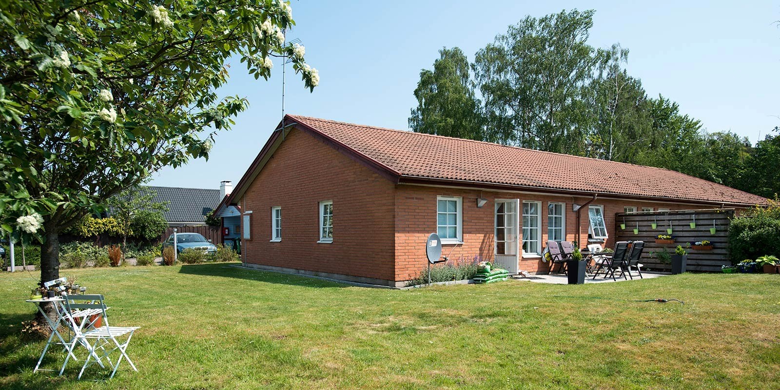 Ett rött tegelhus med brunt tak och en välskött grön gräsmatta i förgrunden. Det finns en vit sittgrupp på gräsmattan och ytterligare utemöbler nära husets entré. Träd och buskar omger fastigheten