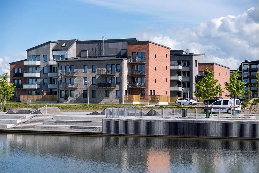 En modern bostadsbyggnad vid Råbysjön med tegel och mörka material, balkonger, parkerade bilar, och reflektion i vattnet. Det är grönområden och unga träd runtomkring.