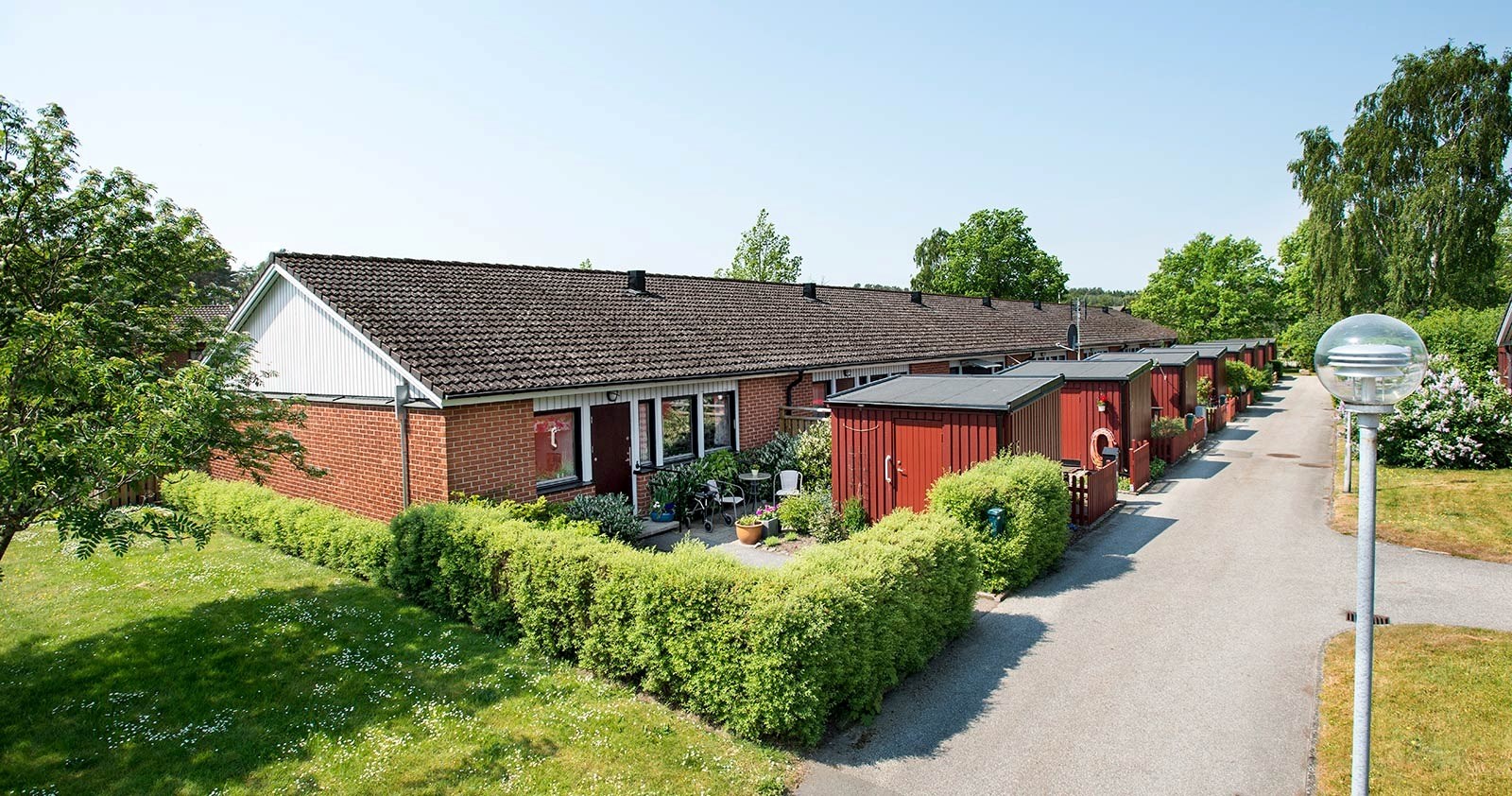 Ett bostadsområde med en radhus. Varje hus är byggt av röda tegelstenar och har ett mörkt tak. Framför husen finns prydligt trimmade gröna buskar. En gångväg löper längs med husraden. Det finns träd i bak- och förgrunden.
